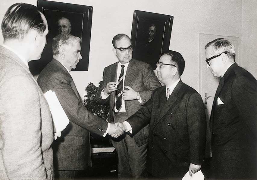 Two people from a group of five shake hands after signing a contract in 1960