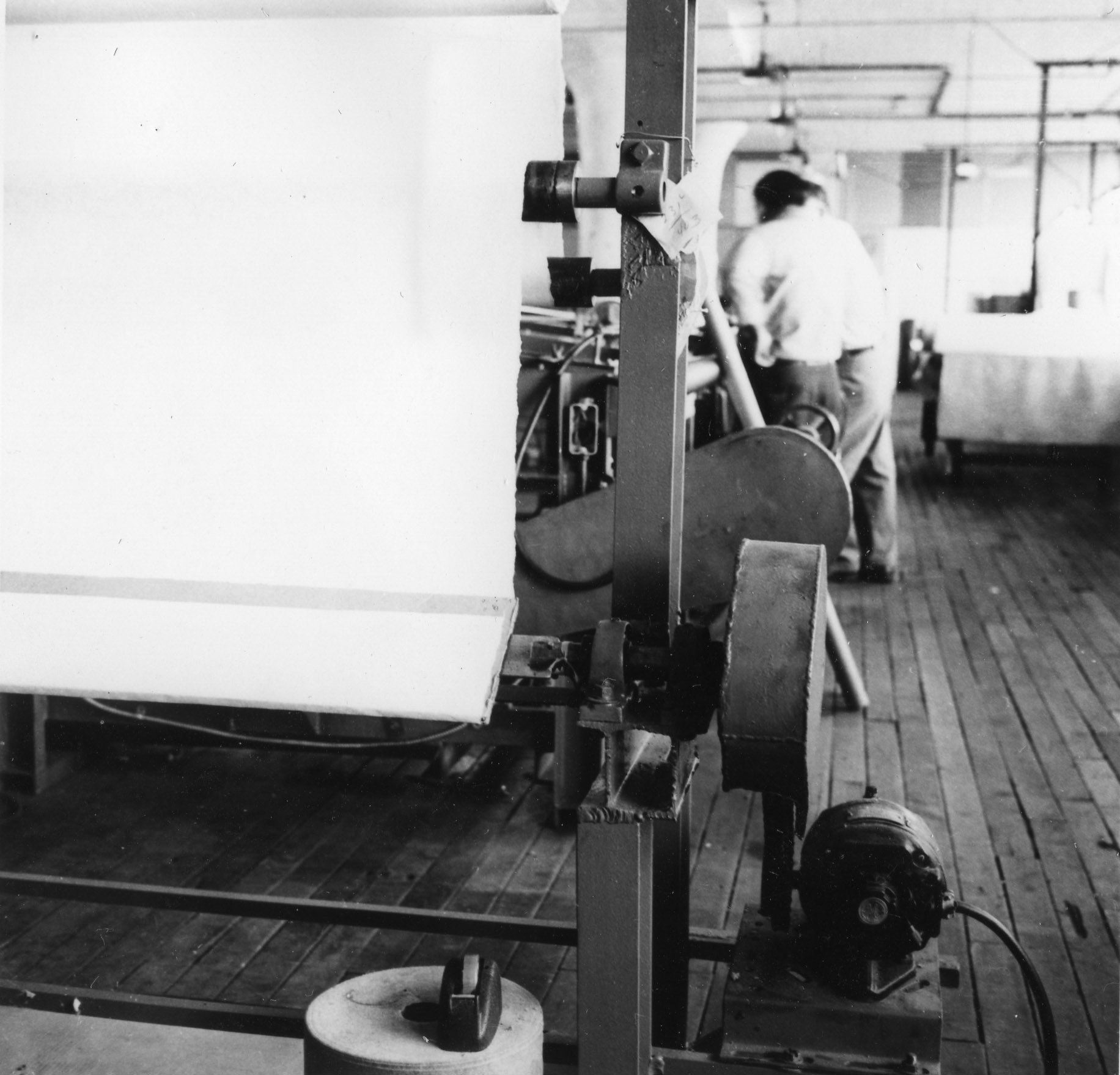 Picture shows an old looking, grey photo of a room with wooden floor and a machine for nonwoven production