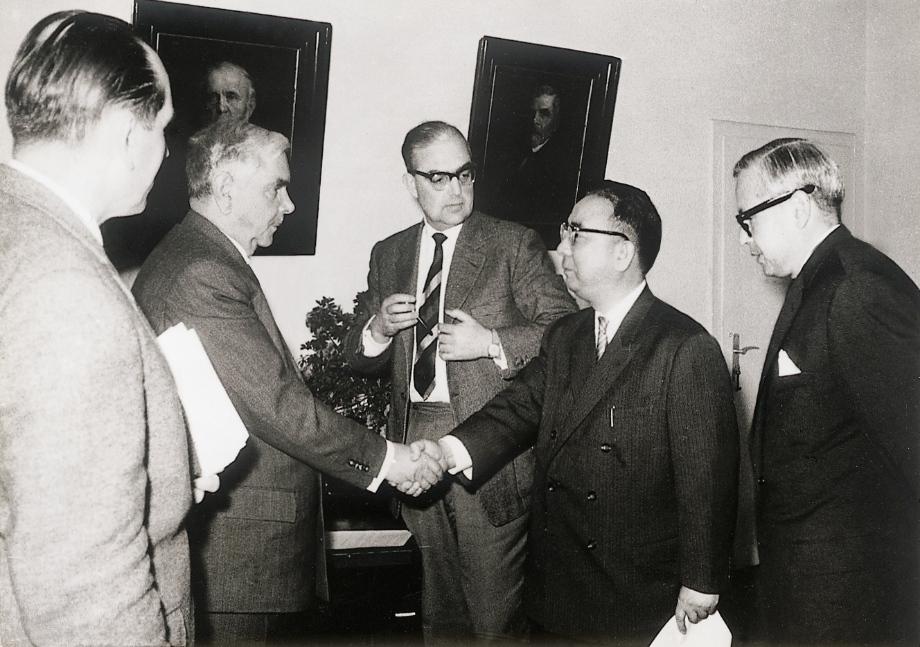 The picture shows five men wearing suits and standing in front of each other. Two of them are shaking hands.