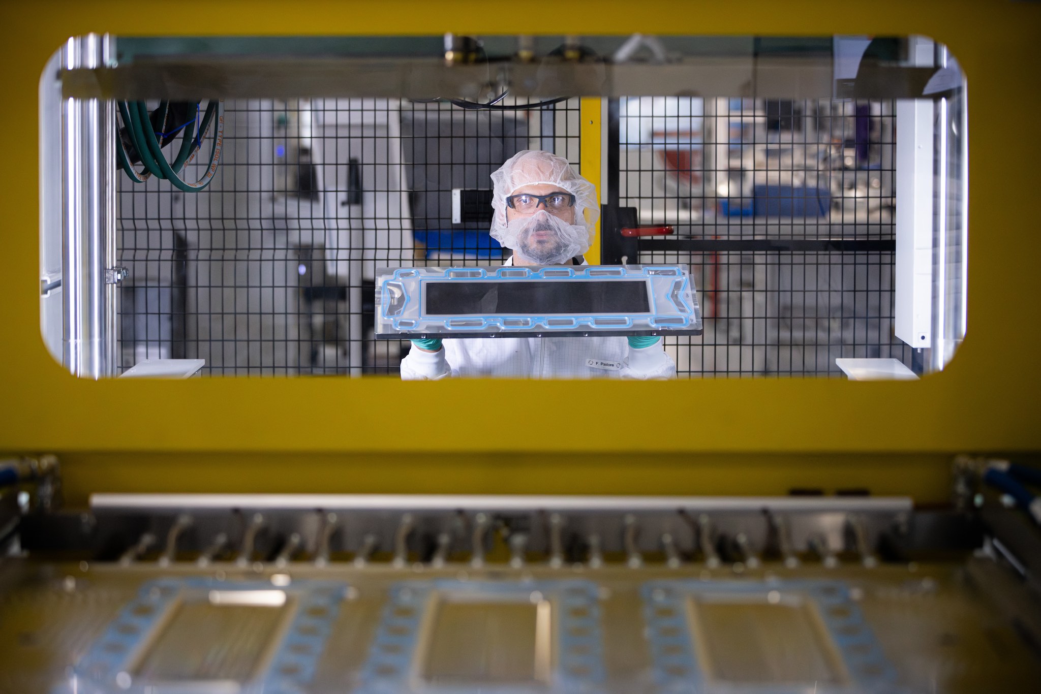 Picture shows a man wearing white laboratory clothing who is looking towards a yellow frame that seems to be a machine that produces fuel cell components