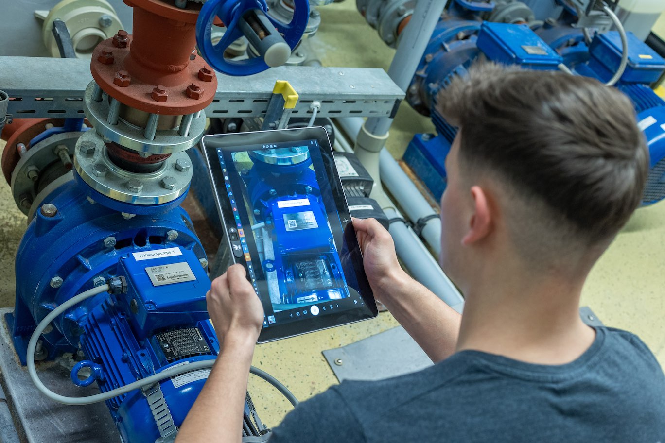 Picture shows the back of a man who is looking at an ipad that the is holding over a blue machine. The ipad screen zooms on the blue machine