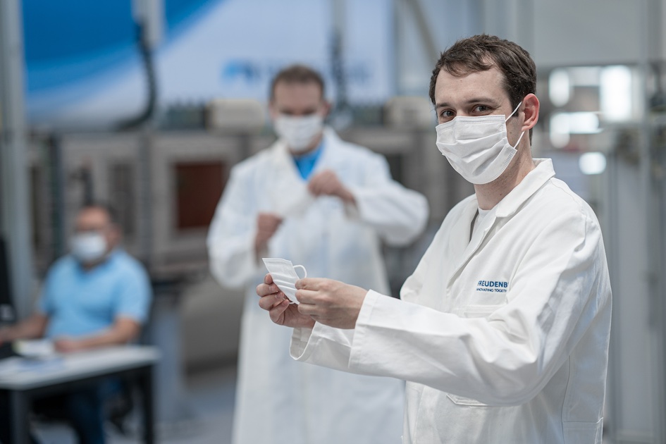 Picture shows a man wearing white laboratory clothing who is wearing a face mask and is holding one in his hands