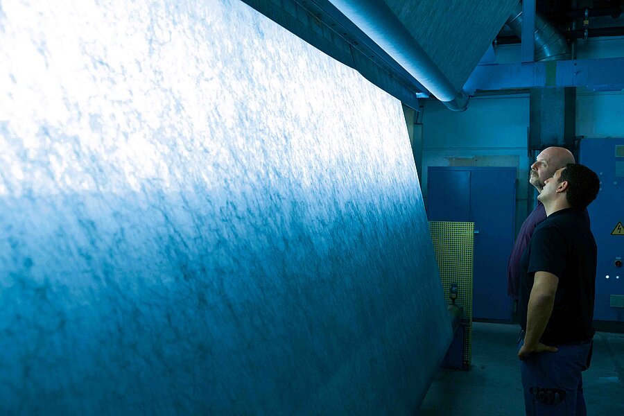 The picture shows two men looking at a large textile roll which visualizes the spunbonded nonwoven production 