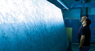 The picture shows two men looking at a large textile roll which visualizes the spunbonded nonwoven production 