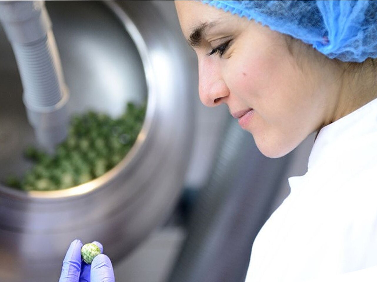 a woman with a hair net and gloves reviewing a confectionery piece in front of a big stell tank with more green confectionery in it