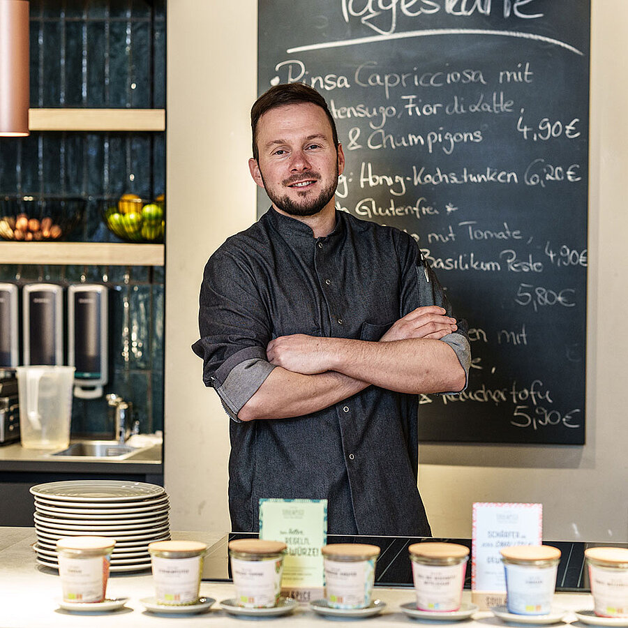 Ein Mann steht hinter einem Tresen in einem Bistro, verschränkt die Arme und schaut freundlich in die Kamera.