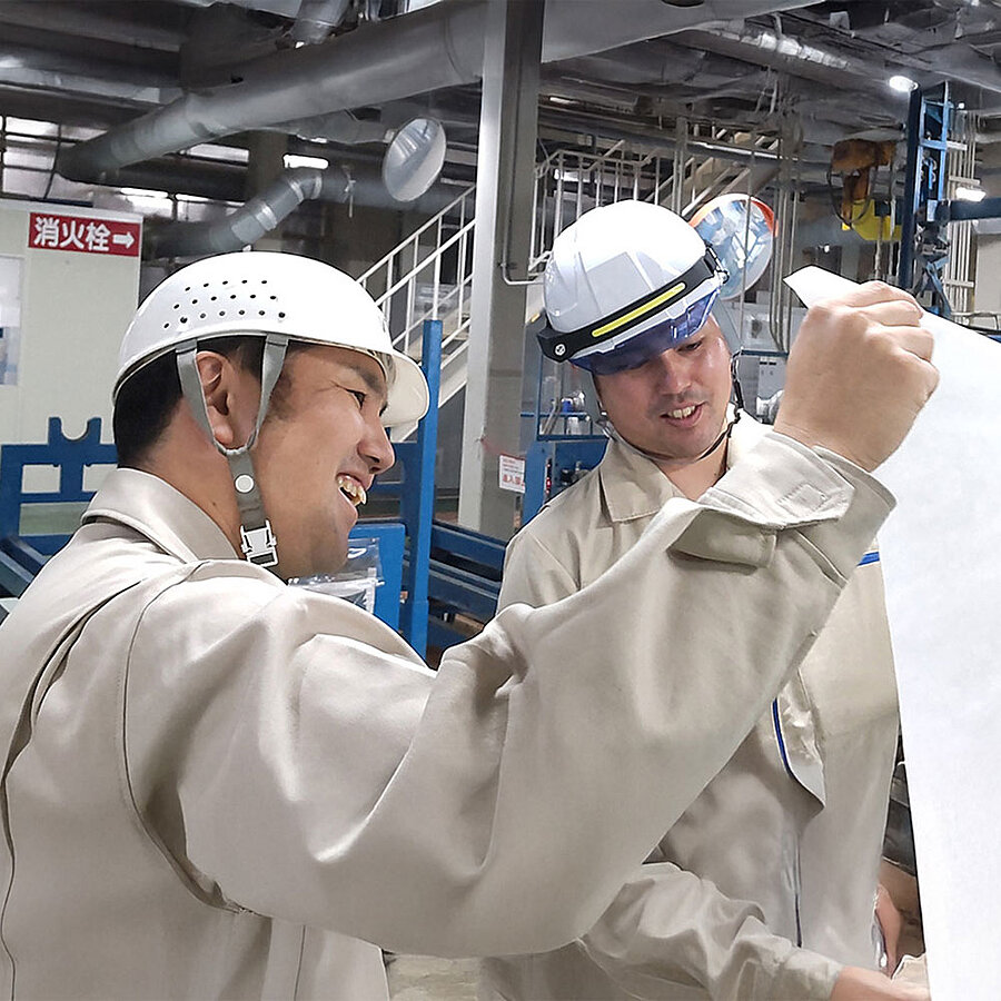 Two people wearing work clothing and white safety helmets inspecting the quality of a nonwoven fabric in a production environment.