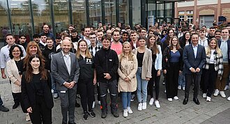 Das Bild zeigt eine große Gruppe von Menschen, die vor einem Gebäude stehen und in die Kamera lächeln
