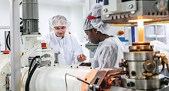 A man and a woman wearing white coats and hairnets stand behind a piece of laboratory equipment, working intently.