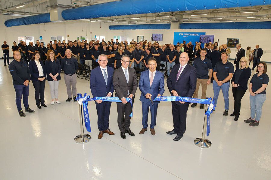 The picture shows four men in a hall cutting a blue ribbon. In the background, many people stand and watch