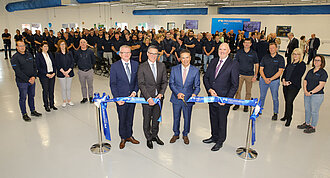 The picture shows four men in a hall cutting a blue ribbon. In the background, many people stand and watch