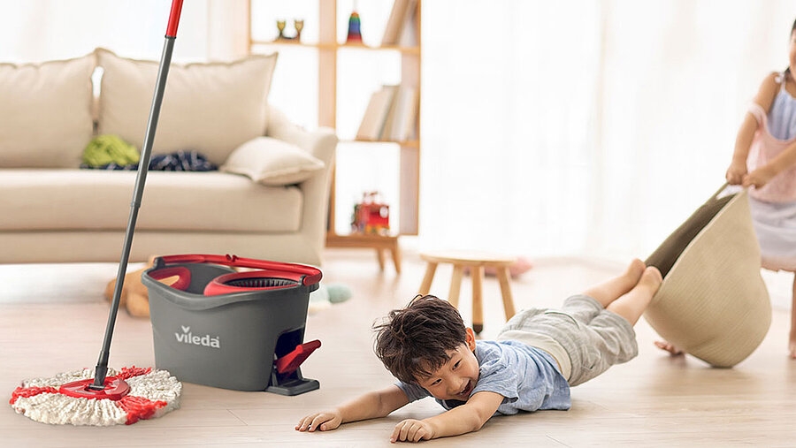 zwei asiatische Kinder tollen auf dem Fußboden im Wohnzimmer. Links neben den Kindern steht ein Putzeimer mit Wischmopp von vileda.