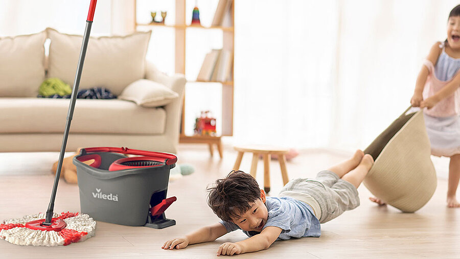 Two Asian children are playing on the floor in the living room. To the left of the children is a cleaning bucket with a Vileda mop.