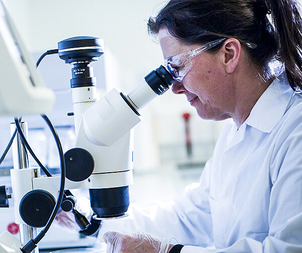 Female scientist wearing a white lab coat and safety glasses is looking into a microscope.