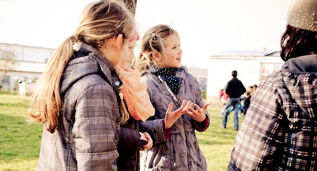 Three children standing next to each other in a park talking to each other