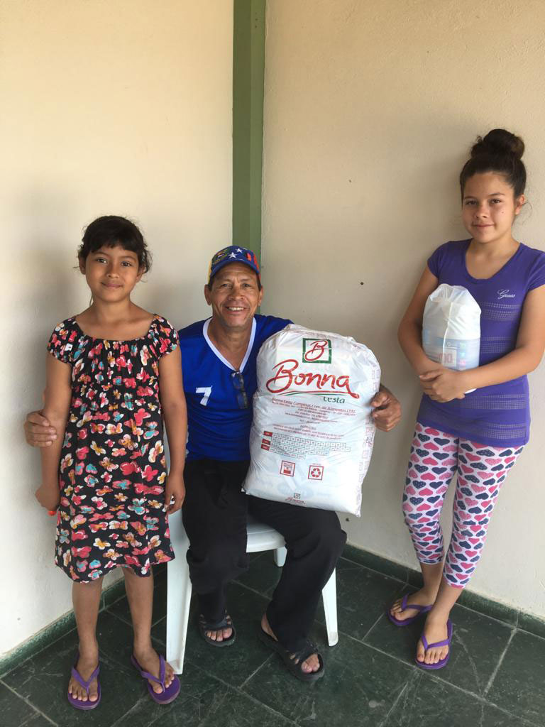 Man sitting on a chair holding a bag of food donations, two children standing next to him.